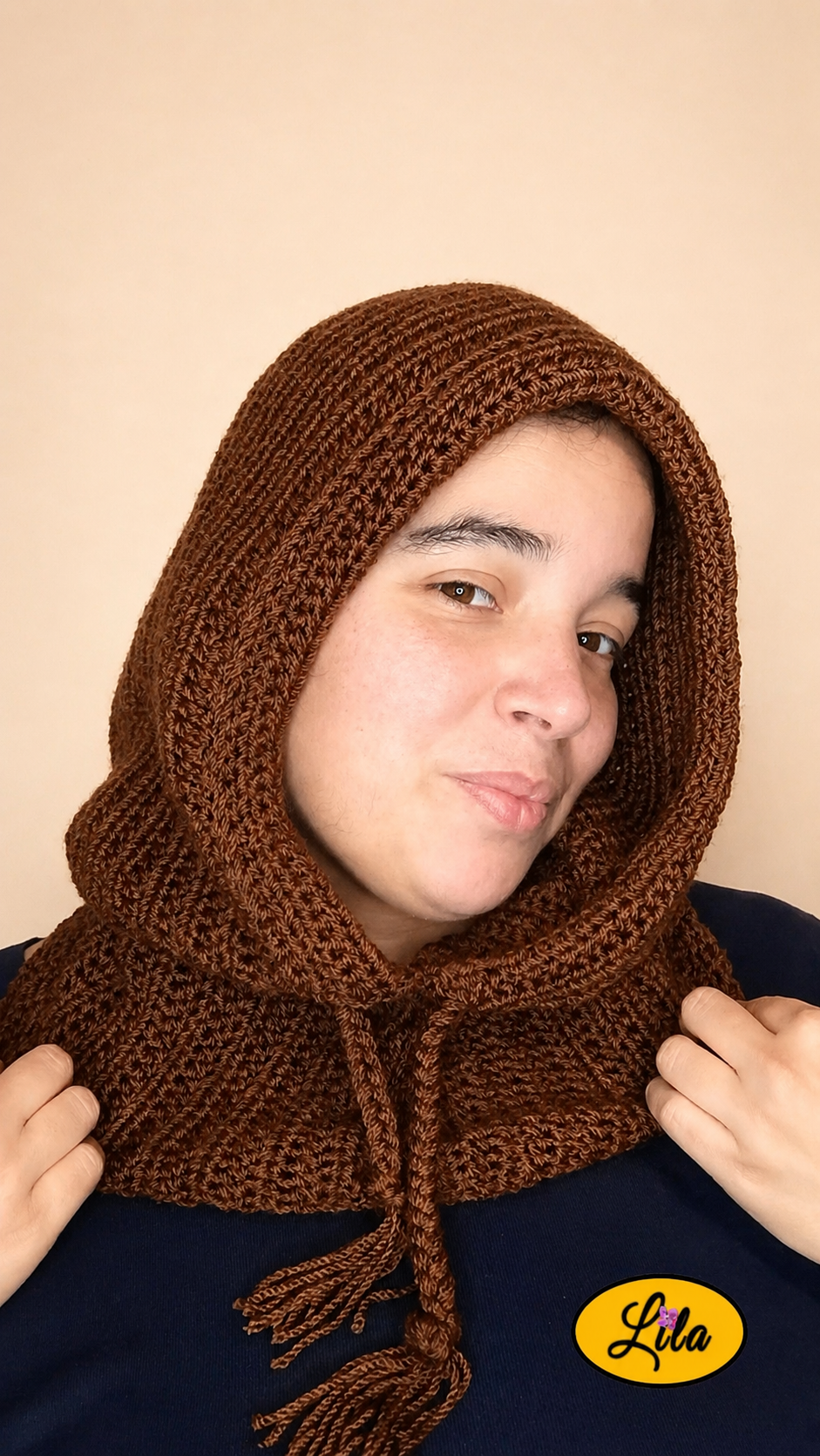 Capuche brune tricotée à la main avec cordons tressés portée par une jeune femme sur fond crème avec col descendant sur le cou tête légèrement inclinée vers la gauche et mains tenant le bas du col style cocooning et élégant