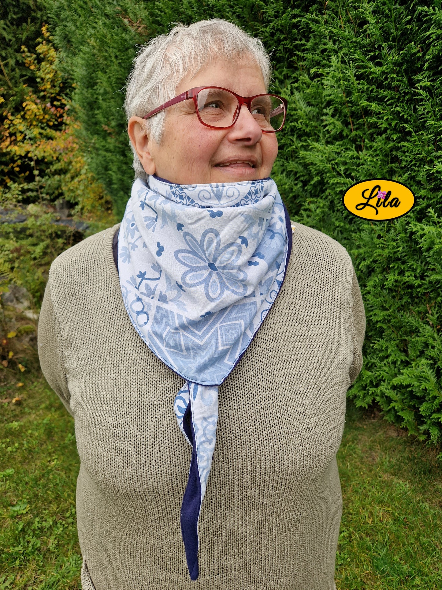 Foulard triangulaire à motifs bleus porté fermé par une dame à lunettes dans un jardin avec nœud caché sous la pointe du foulard femme de face regard tourné vers la gauche avec sourire style élégant et chaleureux