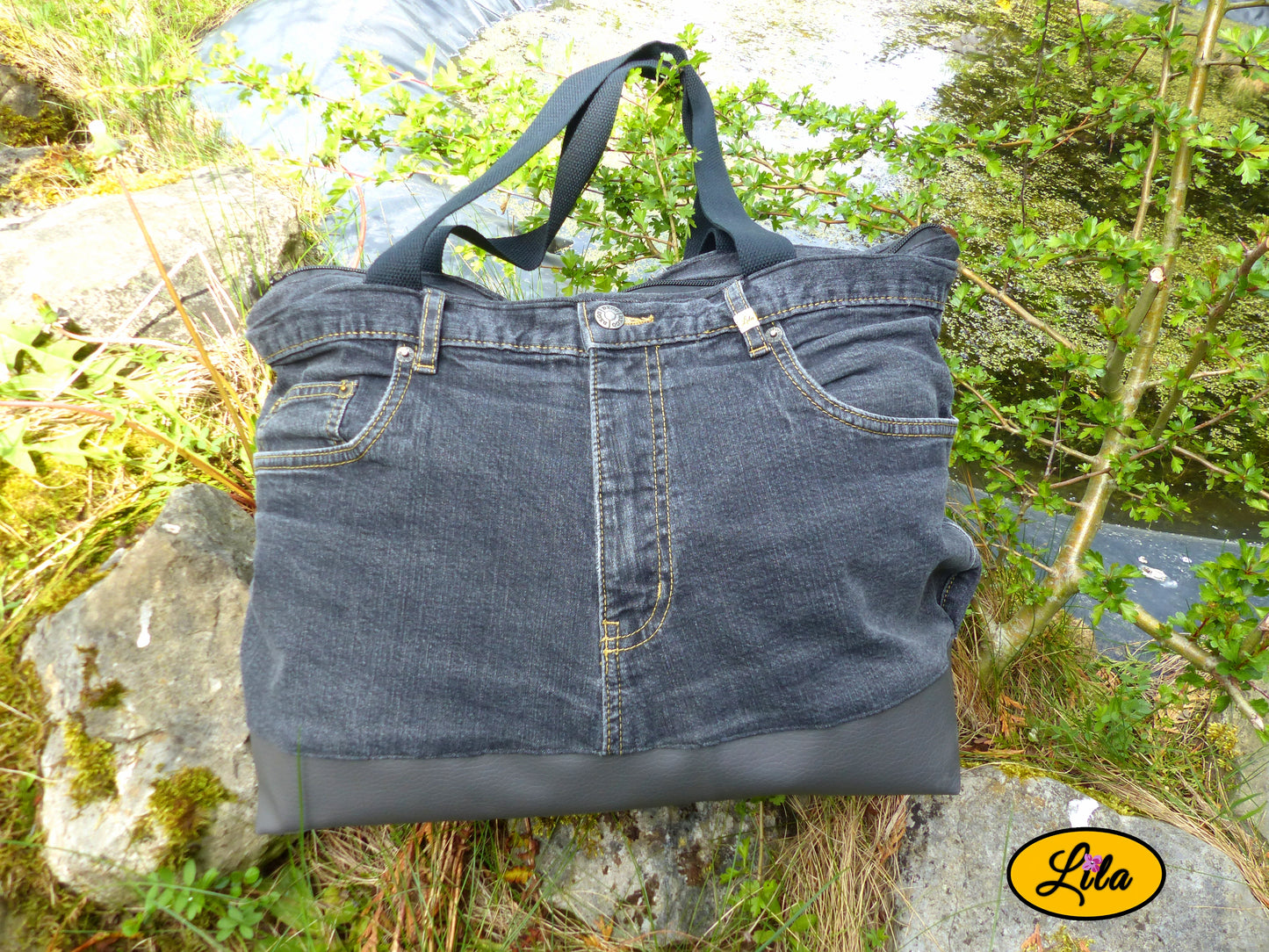 Grand sac en jeans noir recyclé et simili cuir gris foncé posé sur une pierre devant un étang dans un décor naturel mettant en valeur son design artisanal et écoresponsable