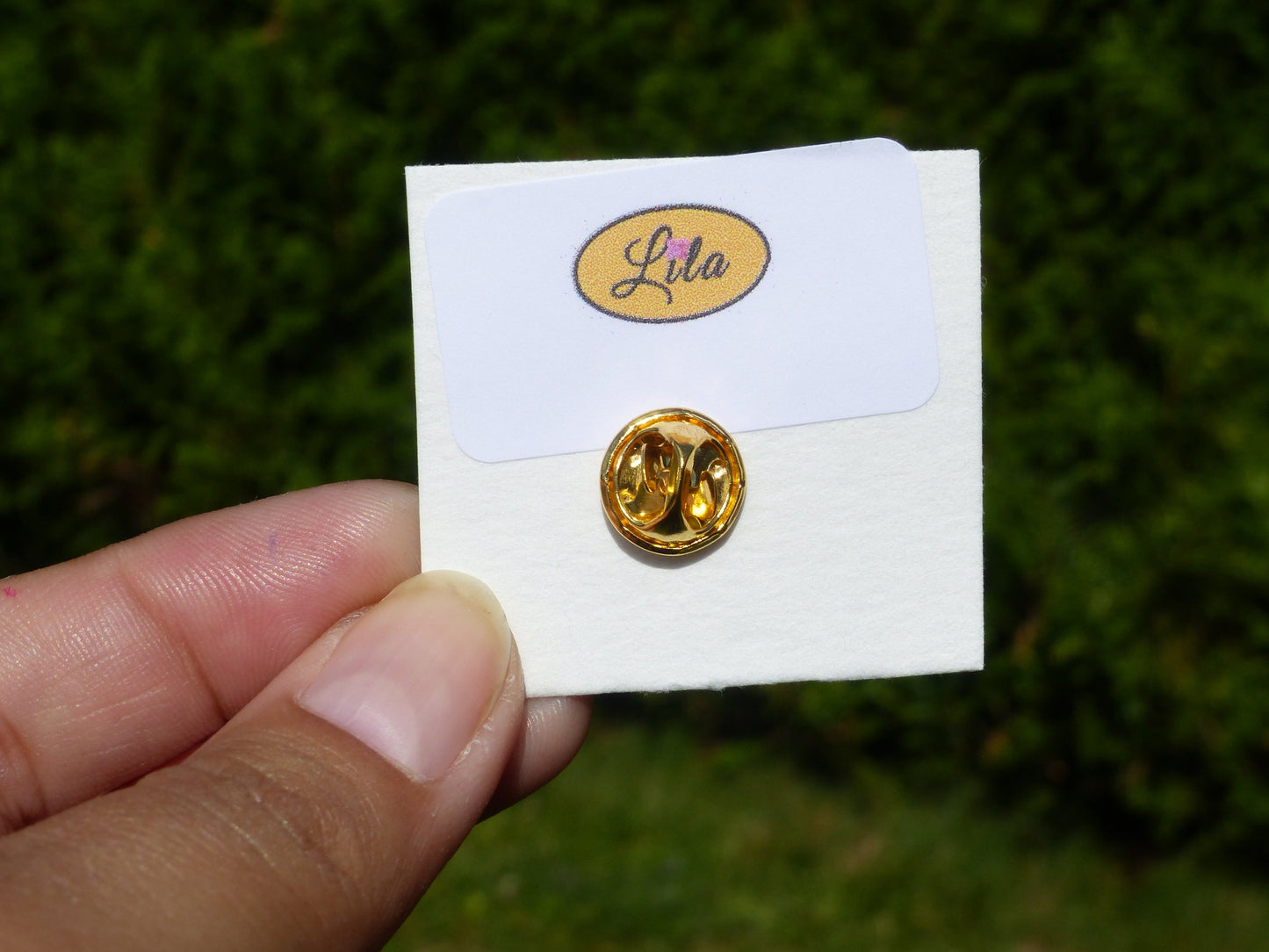 Pin’s bonhomme rose réalisé en pâte polymère avec attache dorée sur carton de présentation blanc photographié devant une haie style kawaii et artisanal