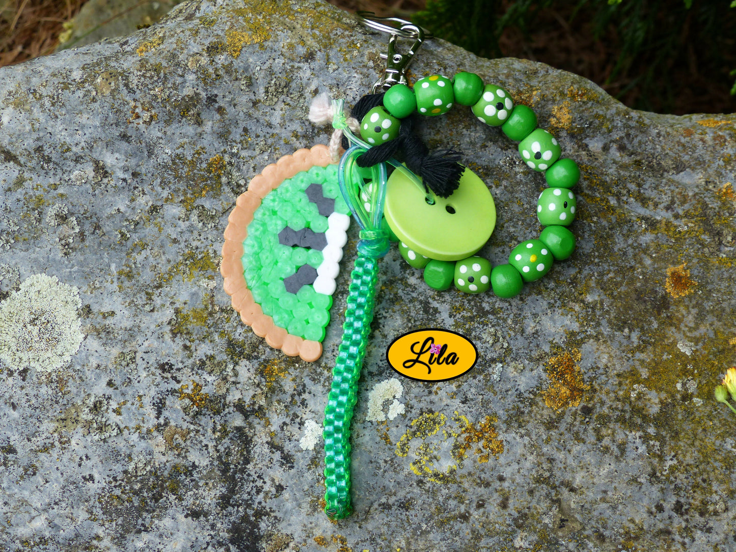 Porte-clés Kiwi vintage posé sur une pierre vue de haut avec demi-tranche de kiwi en perles à repasser, grosses perles de bois vertes, bouton vert recyclé et scoubidou vert visibles côte à côte style récup et kawaii