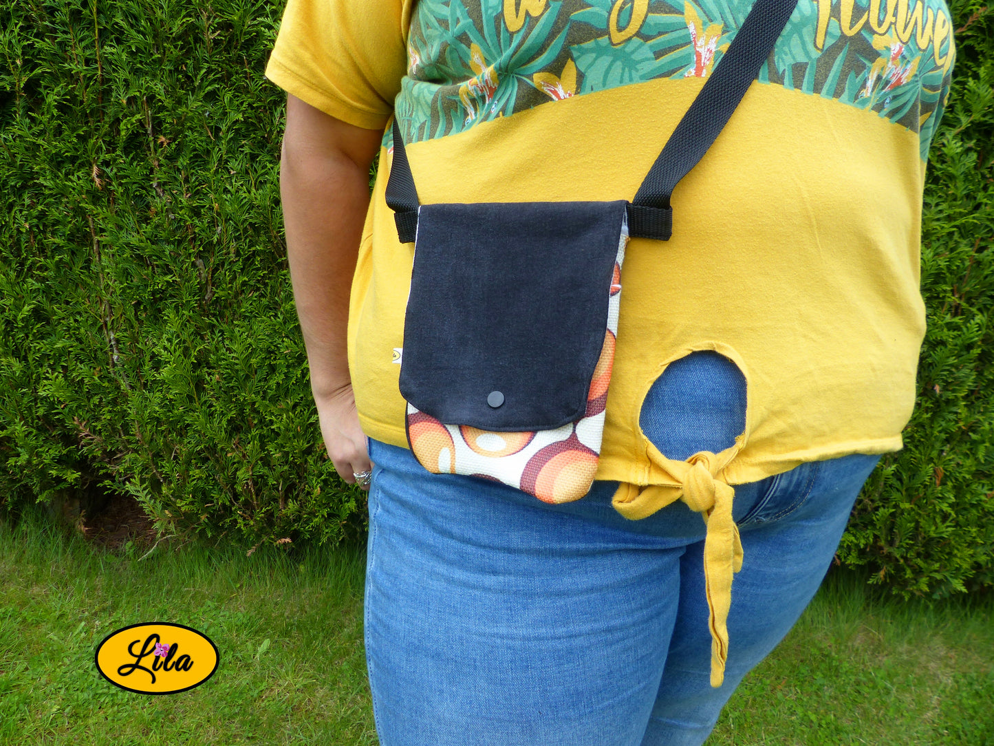 Sac bandoulière pour téléphone en jeans noir avec rabat en ancienne nappe vintage à motifs de ronds orange porté par une jeune femme devant une haie dans un jardin avec photo cadrée sur le sac mettant en valeur son style rétro et artisanal