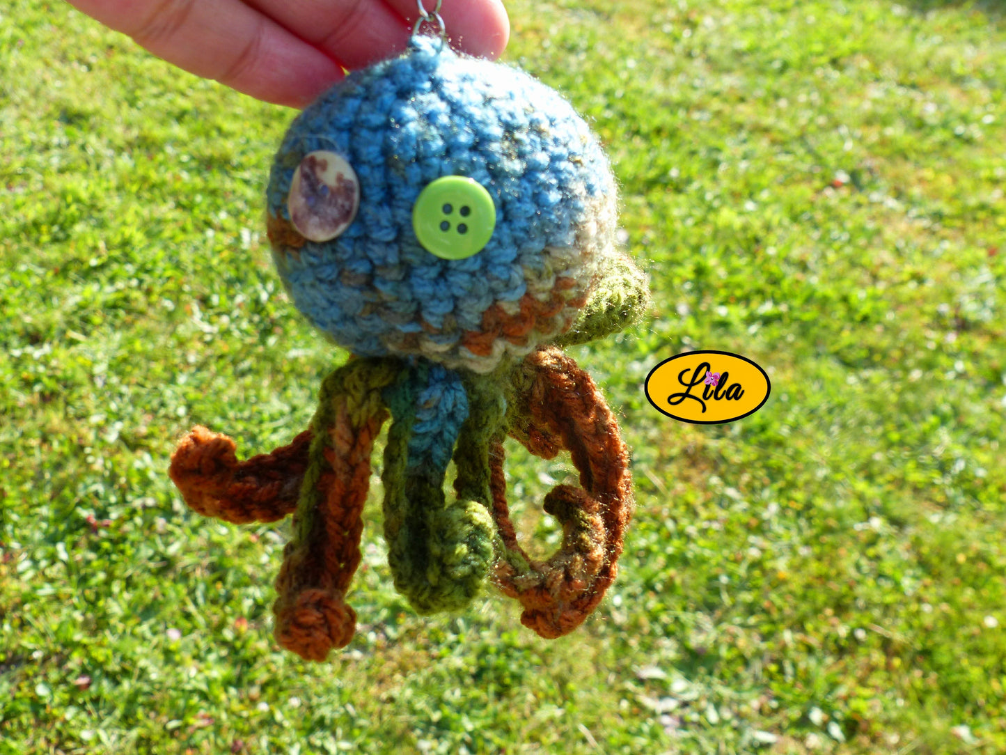 Porte-clé Poulpy pieuvre amigurumi chamarrée aux tons vert bleu et brun réalisée au crochet avec yeux en boutons vert et multicolore tenu dans une main en extérieur dans un jardin style handmade et bohème