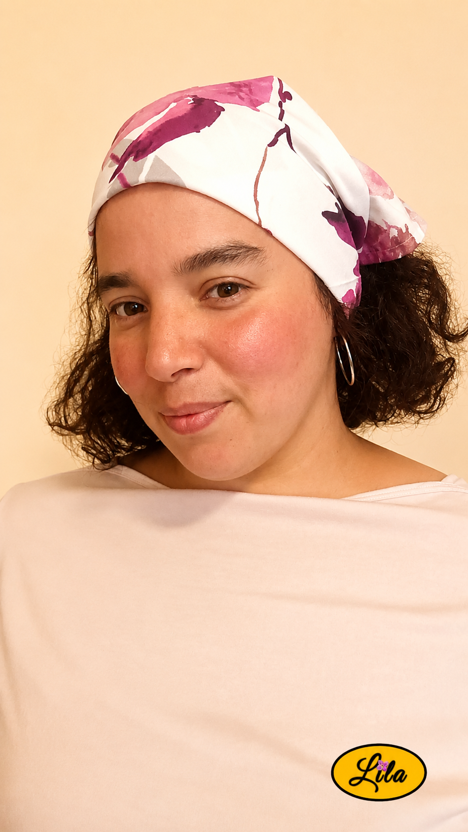 Bandana blanc et gris à motifs de fleurs roses porté dans les cheveux par une femme légèrement de profil gauche avec les cheveux détachés et un léger sourire sur fond crème style romantique et élégant