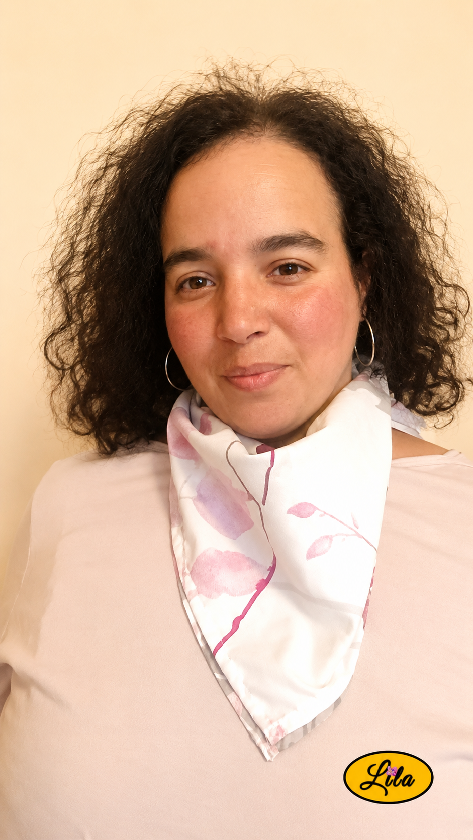 Bandana blanc et gris à motifs de fleurs roses porté autour du cou par une femme de face aux cheveux lâchés sur fond crème style romantique et tendance