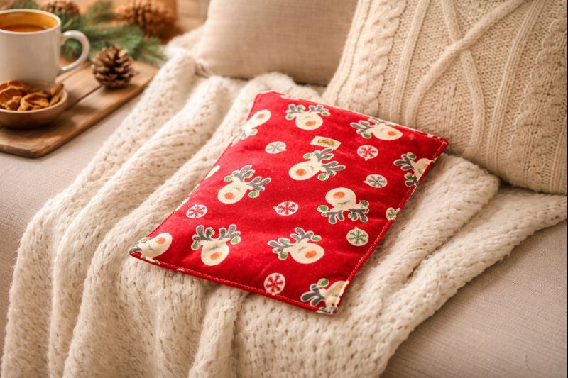 Bouillotte motif rennes sur fond rouge réalisée dans un ancien pyjama recyclé et remplie de noyaux de cerise posée sur un fauteuil avec plaid oreiller et bougie allumée ambiance cocooning et hivernale
