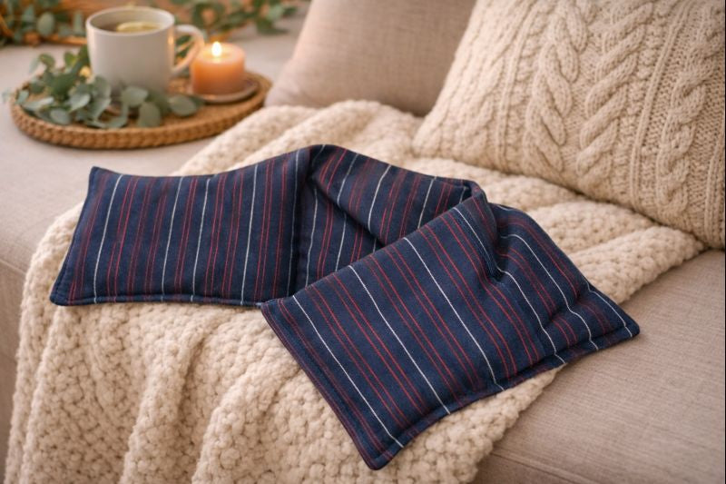 Bouillotte XL bleue à lignes rouges réalisée dans un ancien pyjama recyclé et remplie de noyaux de cerise posée sur un fauteuil avec plaid oreiller et bougie allumée ambiance cocooning et chaleureuse