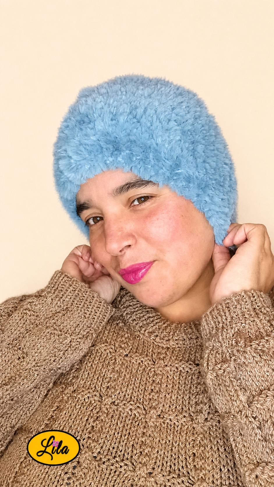 Cache-cou tricoté main bleu effet fausse fourrure porté sur la tête par une femme avec pull brun sur fond crème tête légèrement inclinée à droite et mains tenant le cache-cou style doux et hivernal