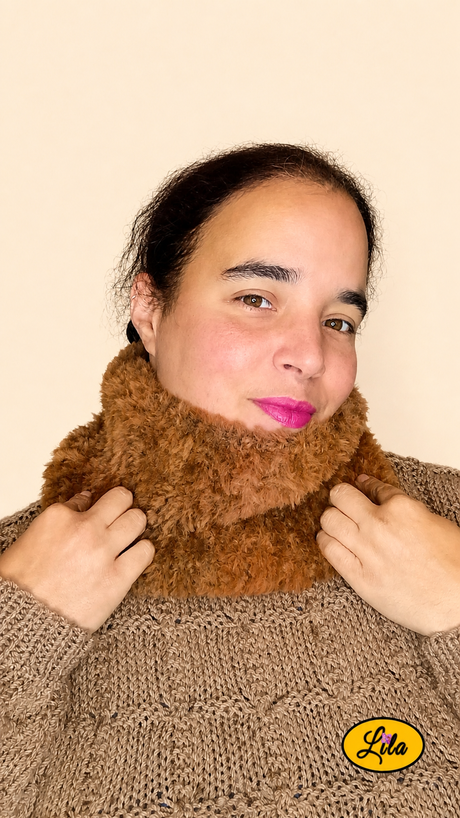 Cache-cou tricoté main brun effet fausse fourrure porté dans le cou par une femme avec pull brun sur fond crème tête légèrement inclinée à gauche et mains tenant le cache-cou style cocooning et élégant