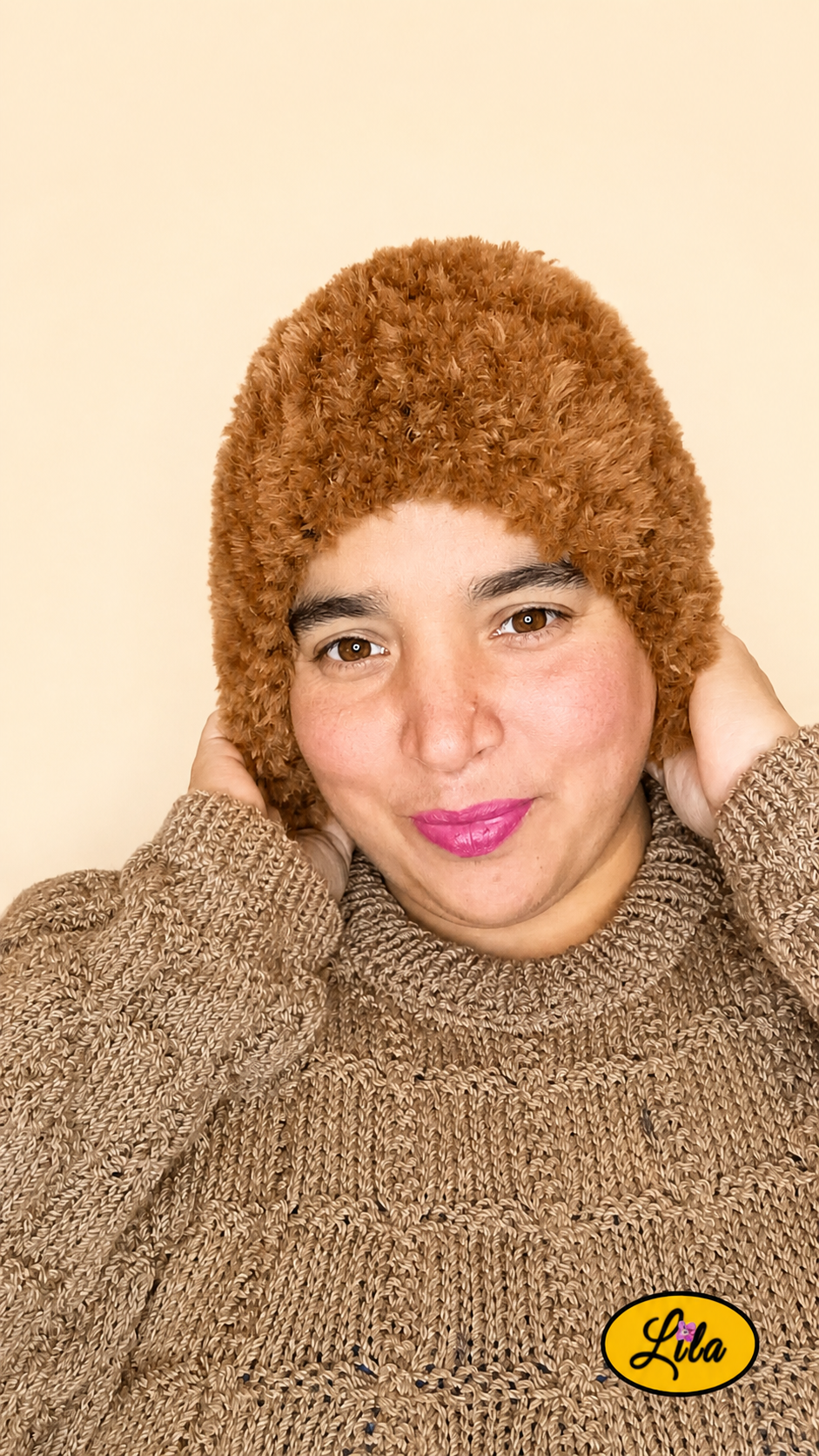 Cache-cou tricoté main brun effet fausse fourrure porté sur la tête par une femme avec pull brun sur fond crème mains posées sur le cache-cou style hivernal et chaleureux
