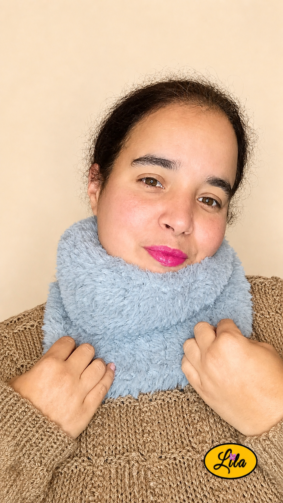 Cache-cou tricoté main gris effet fausse fourrure porté dans le cou par une femme avec pull brun sur fond crème tête penchée à gauche et mains tenant le bas du cache-cou style chaleureux et élégant