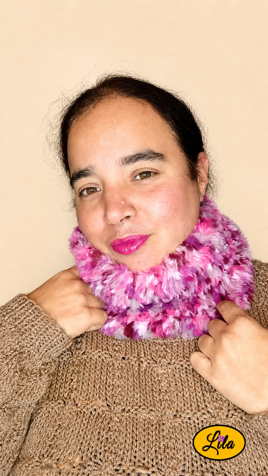 Cache-cou tricoté main rose chamarré effet fausse fourrure porté par une femme avec pull brun sur fond crème tête tournée vers la droite et main tenant le bas du cache-cou style cocooning et féminin