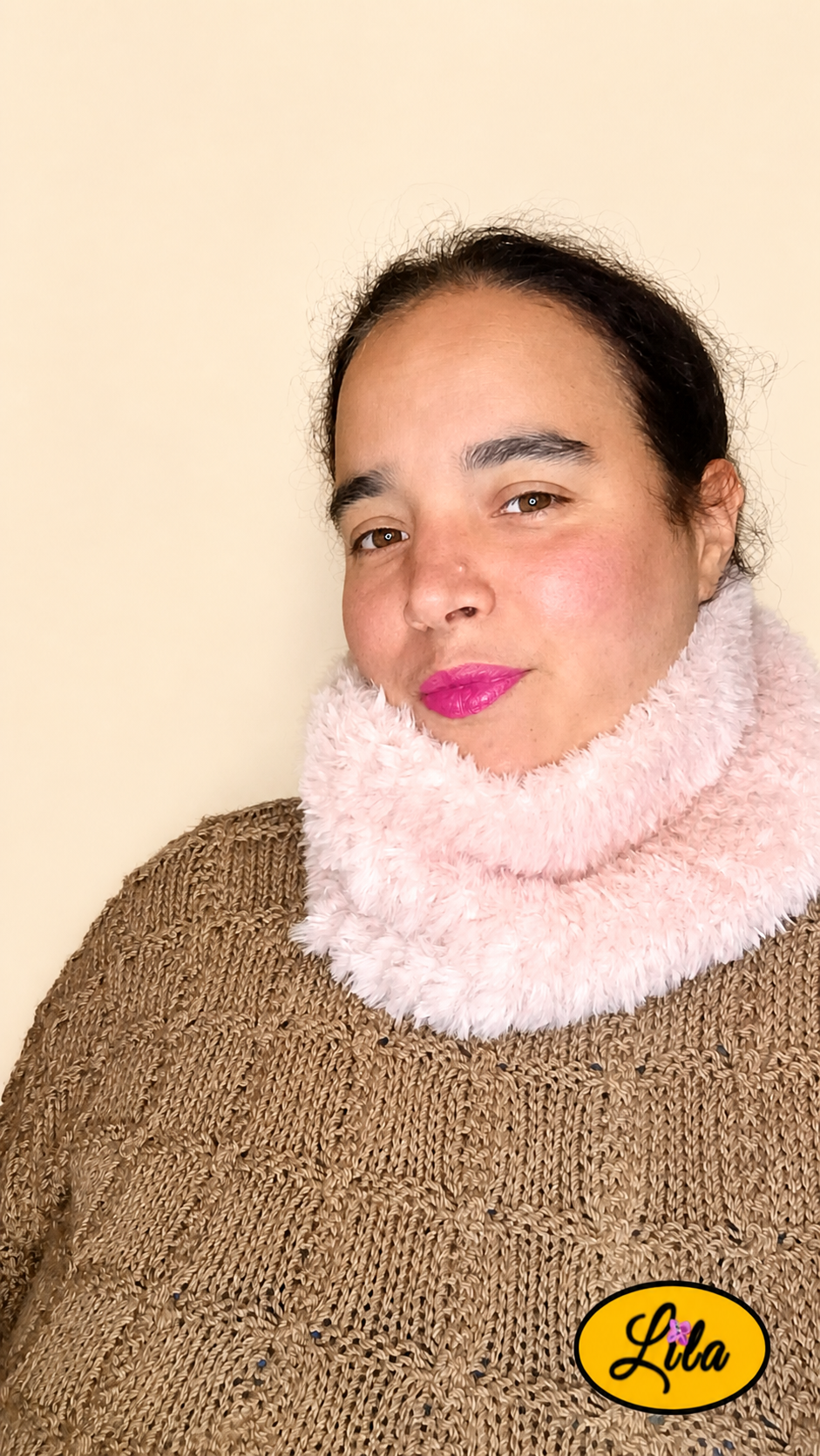 Cache-cou tricoté main rose clair effet fausse fourrure porté dans le cou par une femme avec pull brun sur fond crème tête légèrement tournée vers la droite style féminin et hivernal