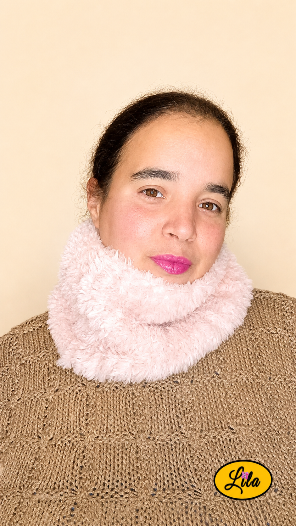 Cache-cou tricoté main rose clair effet fausse fourrure porté dans le cou par une femme avec pull brun sur fond crème tête légèrement tournée vers la gauche style chaleureux et élégant