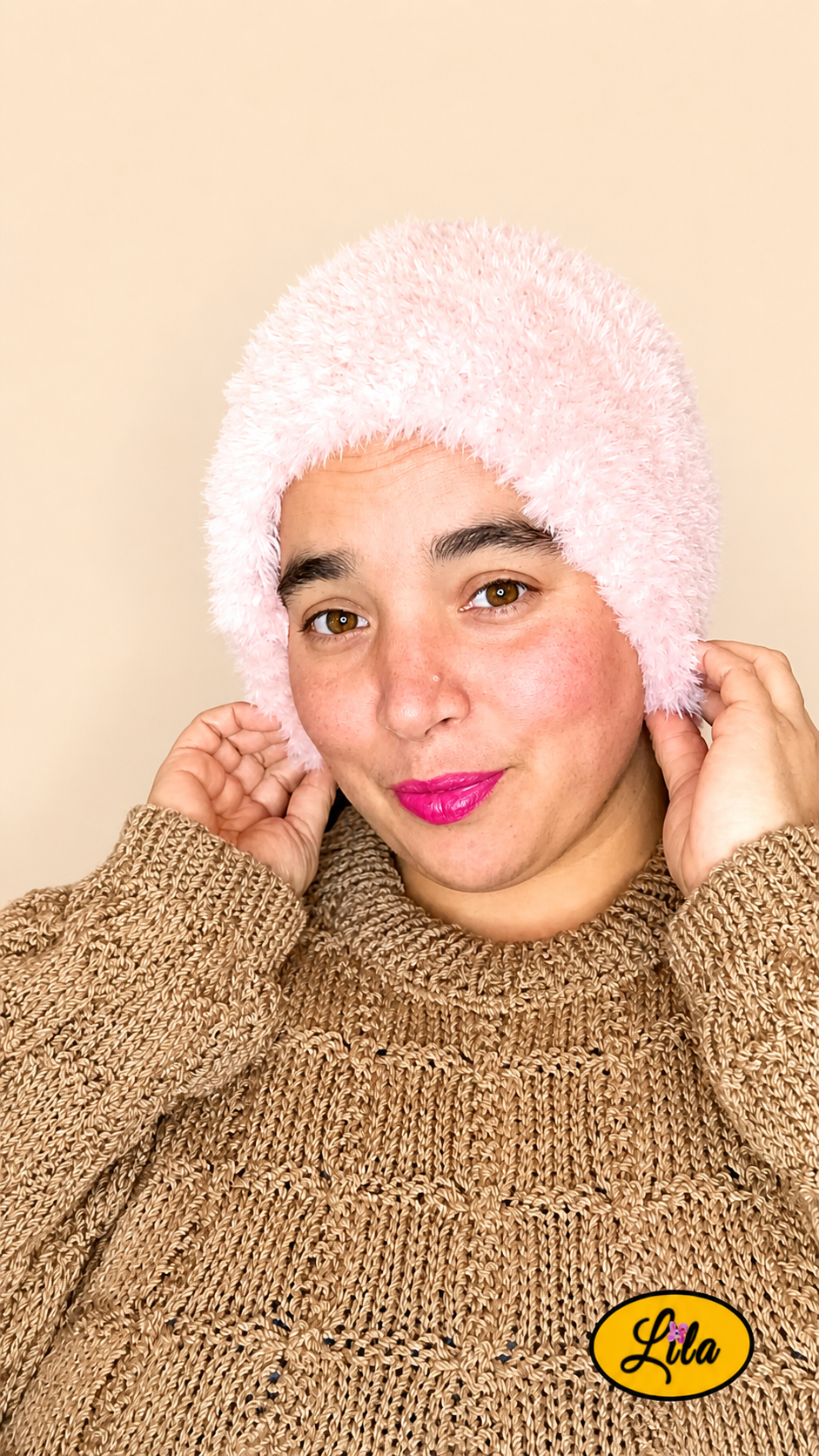 Cache-cou tricoté main rose clair effet fausse fourrure porté sur la tête par une femme avec pull brun sur fond crème tête légèrement tournée vers la droite et mains tenant le bas du cache-cou style doux et cocooning