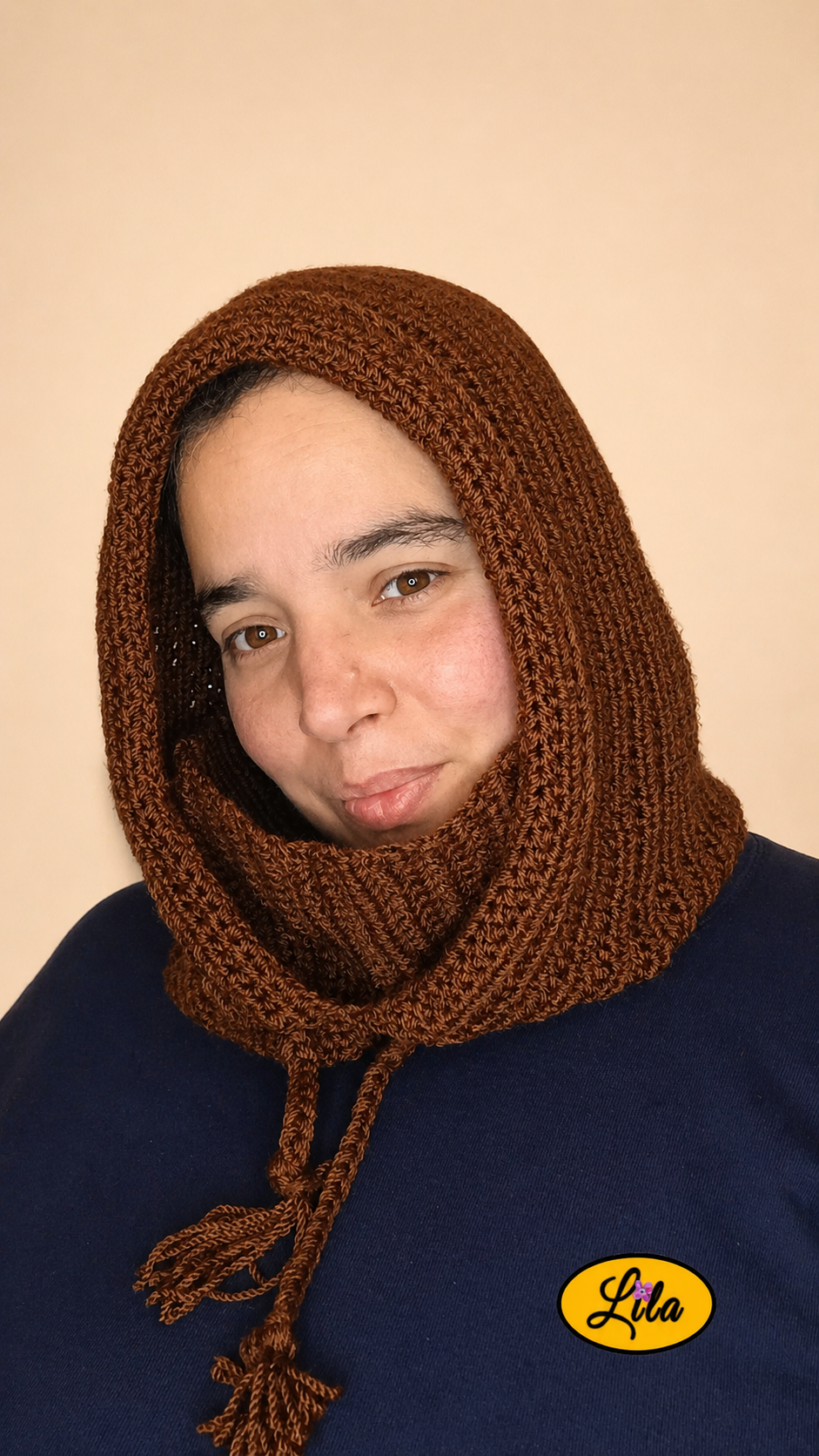 Capuche brune tricotée à la main avec cordons tressés portée par une jeune femme sur fond crème col plié vers l’intérieur tête légèrement inclinée vers la droite style cocooning et hivernal