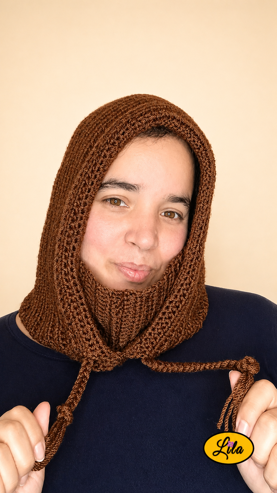 Capuche brune tricotée à la main avec cordons tressés portée par une jeune femme sur fond crème avec col rentré vers l’intérieur tenant les cordons de la capuche dans les mains style hivernal et artisanal