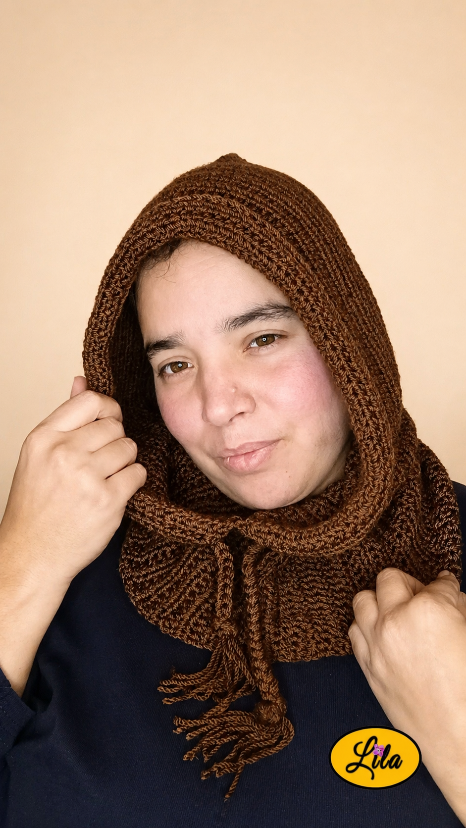 Capuche brune tricotée à la main avec cordons tressés portée par une jeune femme sur fond crème avec col placé dans le cou main gauche sur le bas du col et main droite sur la capuche style chaleureux et cocooning