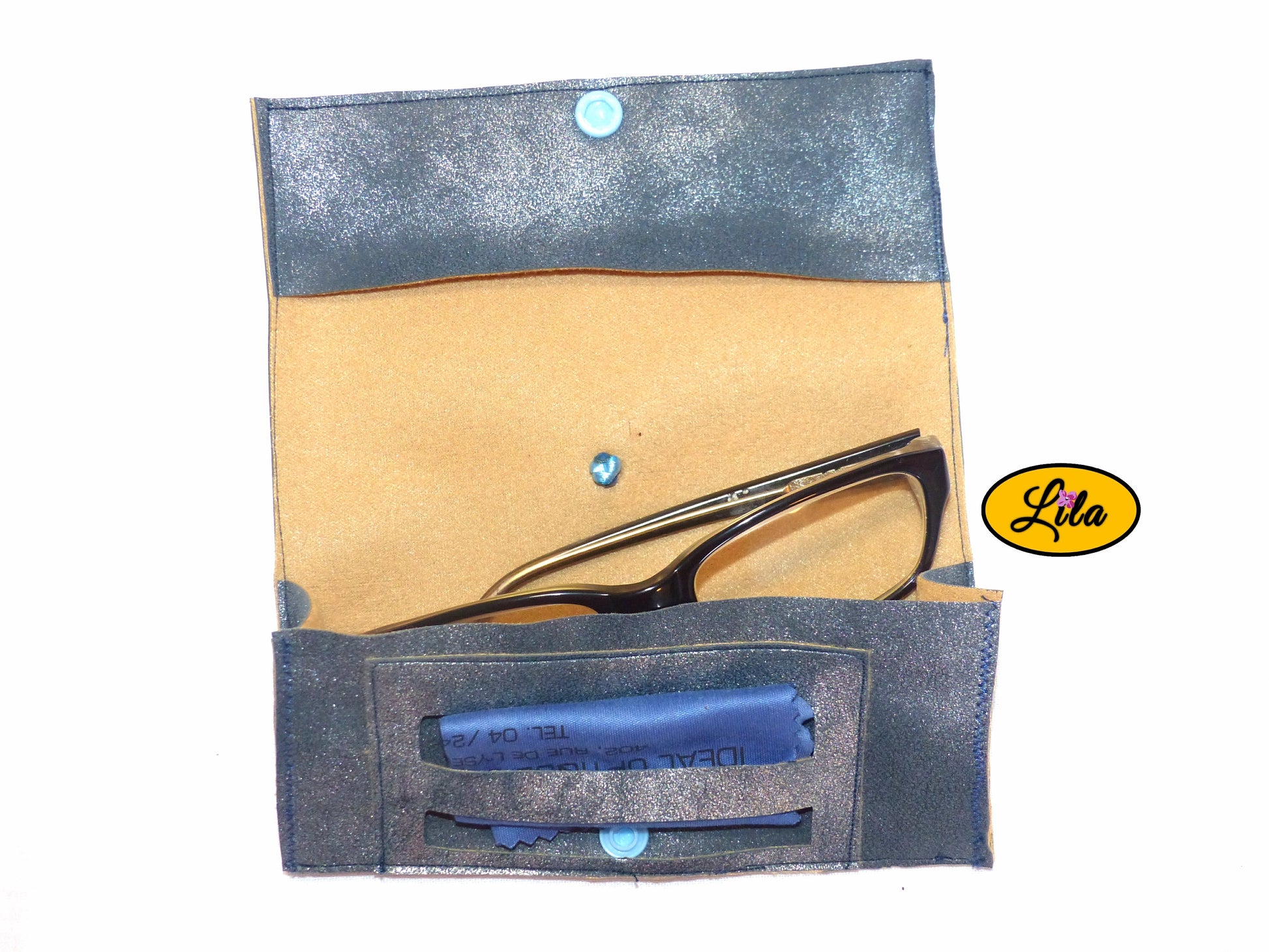 Étui à lunettes en simili bleu argenté ouvert à plat avec paire de lunettes et lingettes rangées dans le compartiment intérieur sur fond blanc style artisanal et fonctionnel