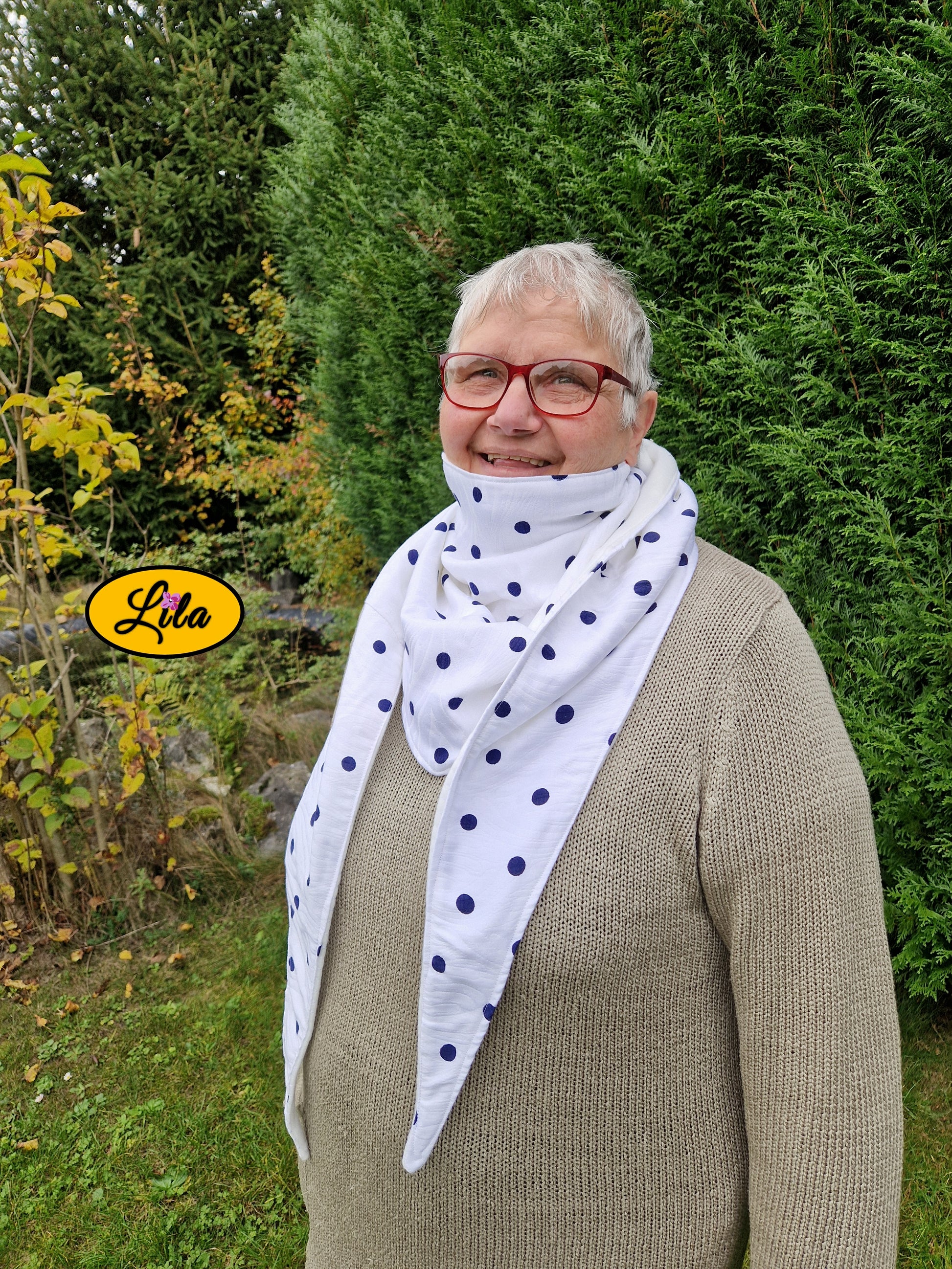 Foulard triangulaire blanc à pois bleu marine porté par une dame à lunettes souriante devant des sapins dans un jardin style cocooning et hivernal