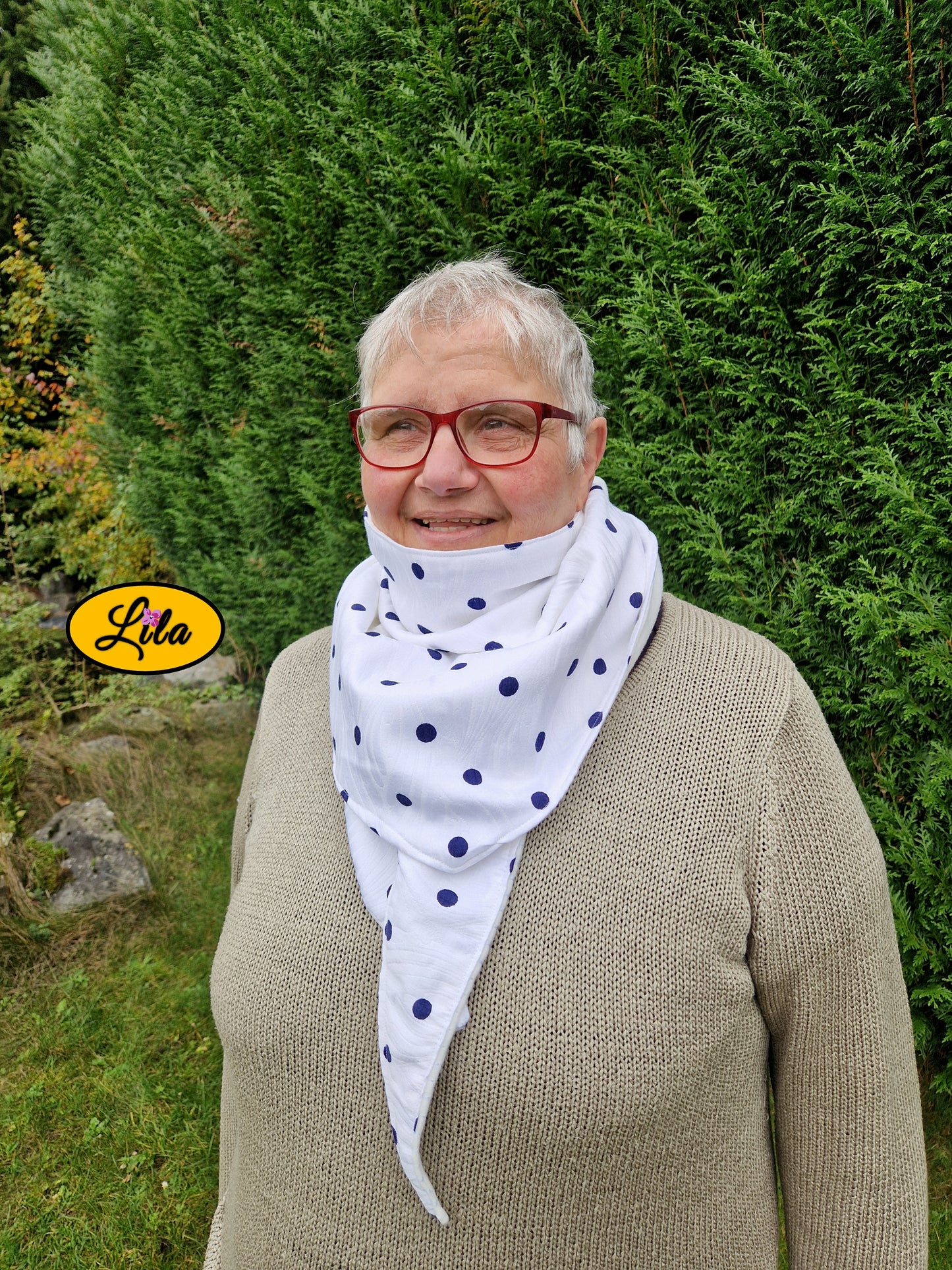 Foulard triangulaire blanc à pois bleu marine porté fermé par une dame avec nœud dissimulé sous la pointe du foulard devant des sapins style rétro et élégant