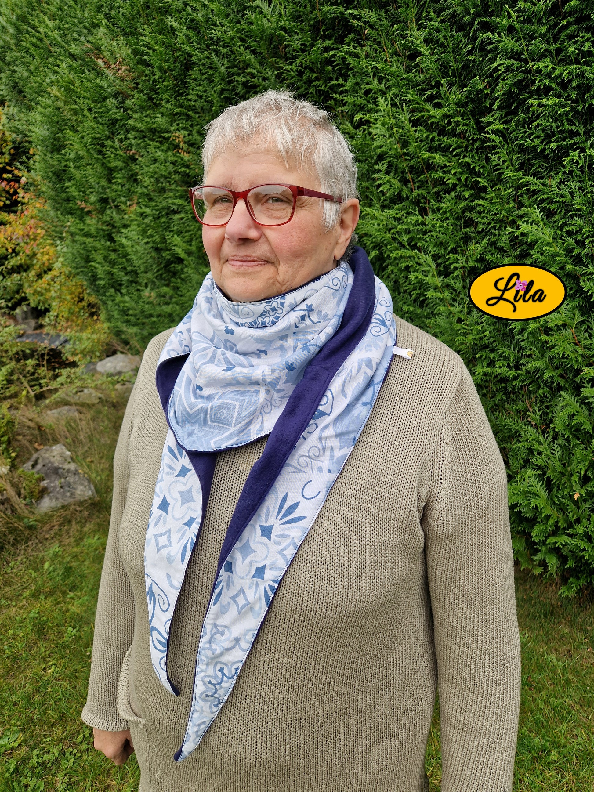 Foulard triangulaire à motifs bleus porté par une dame à lunettes devant une haie de sapins dans un jardin profil droit style hivernal et cocooning