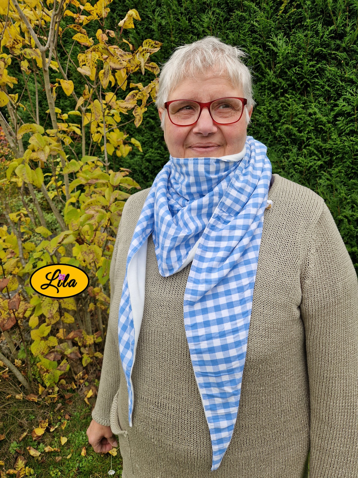 Foulard triangulaire rétro en tissu vichy bleu et blanc porté par une dame à lunettes devant un arbre aux feuilles jaunes regard tourné vers la gauche style automnal et cocooning