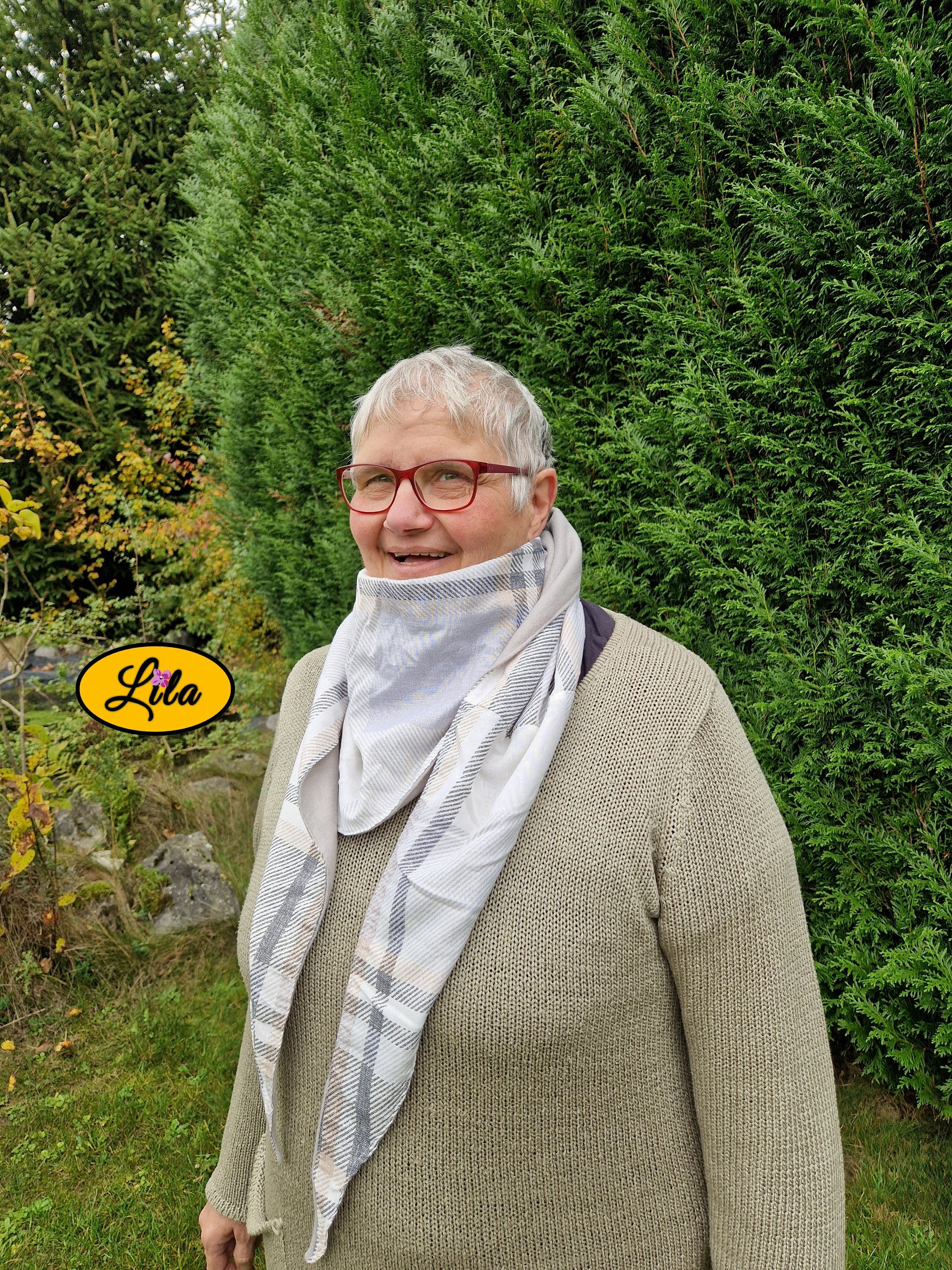 Foulard triangulaire vintage à lignes brun et gris porté par une dame à lunettes dans un jardin automnal près d’une haie profil gauche avec sourire style cocooning et rétro