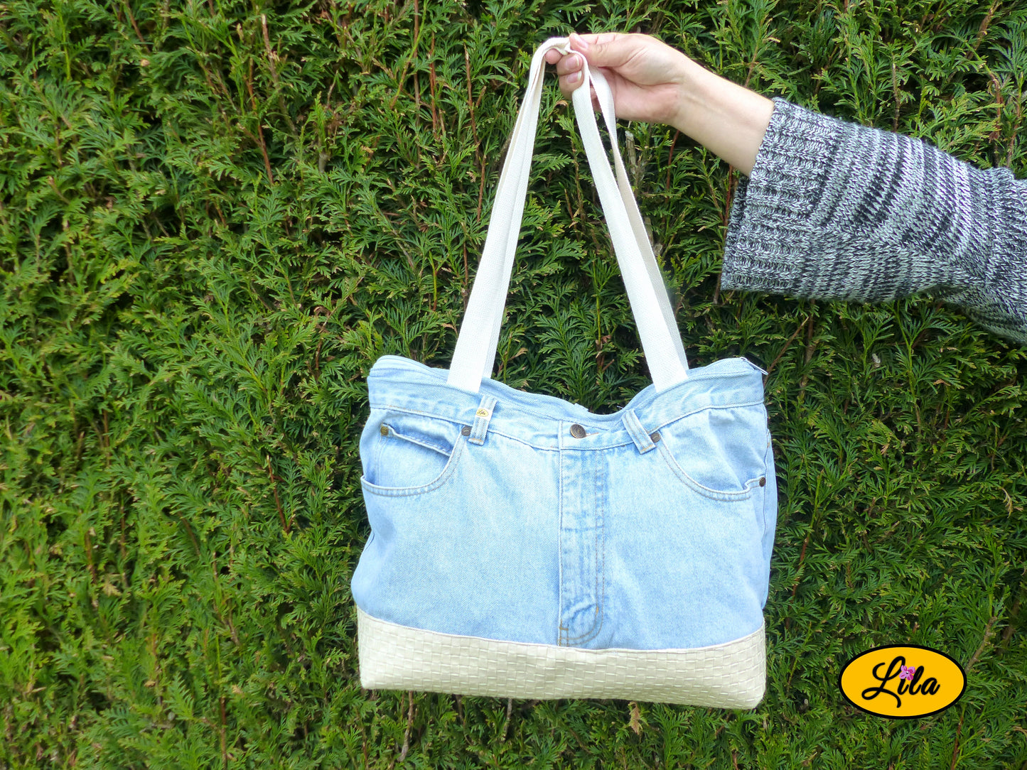 Grand sac en jeans clair recyclé et simili cuir crème tenu à bout de bras devant une haie mettant en valeur son format et son style artisanal