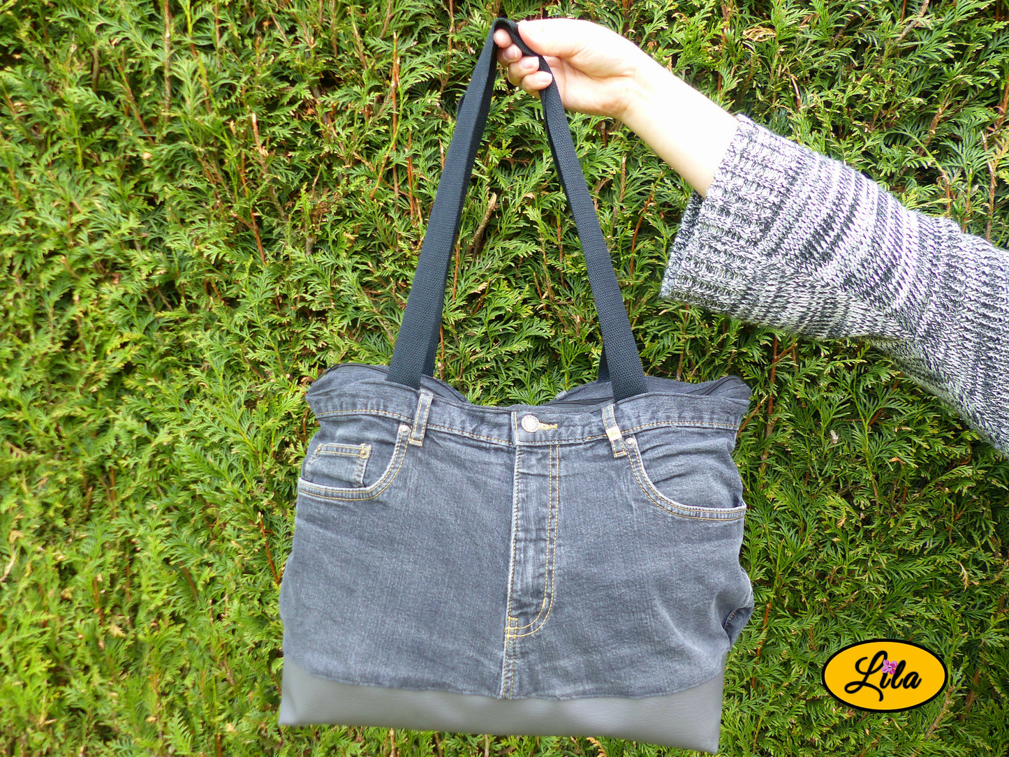 Grand sac en jeans noir recyclé et simili cuir gris foncé porté à bout de bras devant une haie mettant en valeur son format et son style urbain
