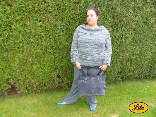 Grand sac en jeans noir recyclé et simili cuir gris foncé tenu à la main par ses anses devant les jambes d'une femme devant une haie mettant en valeur son format et son porté élégant