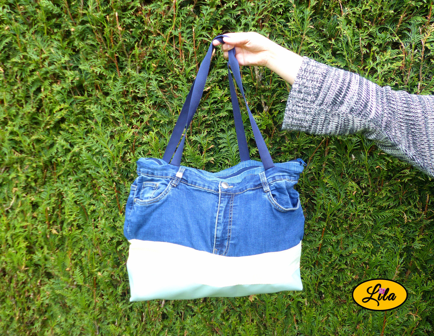 Grand sac en jeans recyclé et simili cuir vert menthe tenu à bout de bras devant une haie mettant en valeur son format et son style artisanal