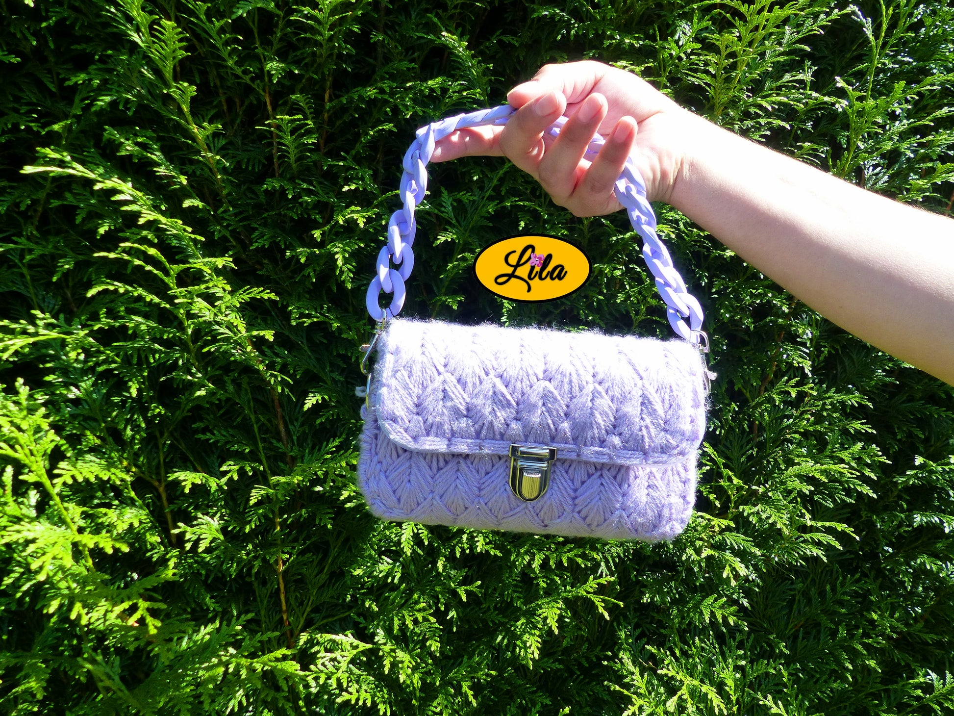 Petit sac à main aux brins lilas sur canevas rigide porté à bout de bras devant une haie style doux et moderne