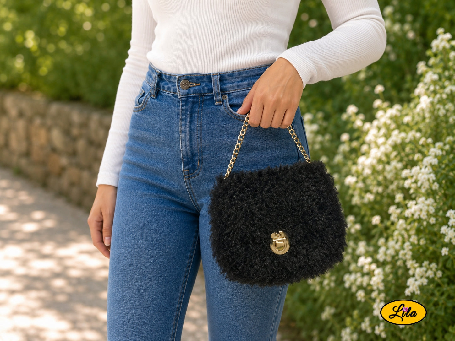 Petit sac à main effet fausse fourrure noire sur canevas rigide porté à la main par un mannequin dans un jardin style chic et tendance