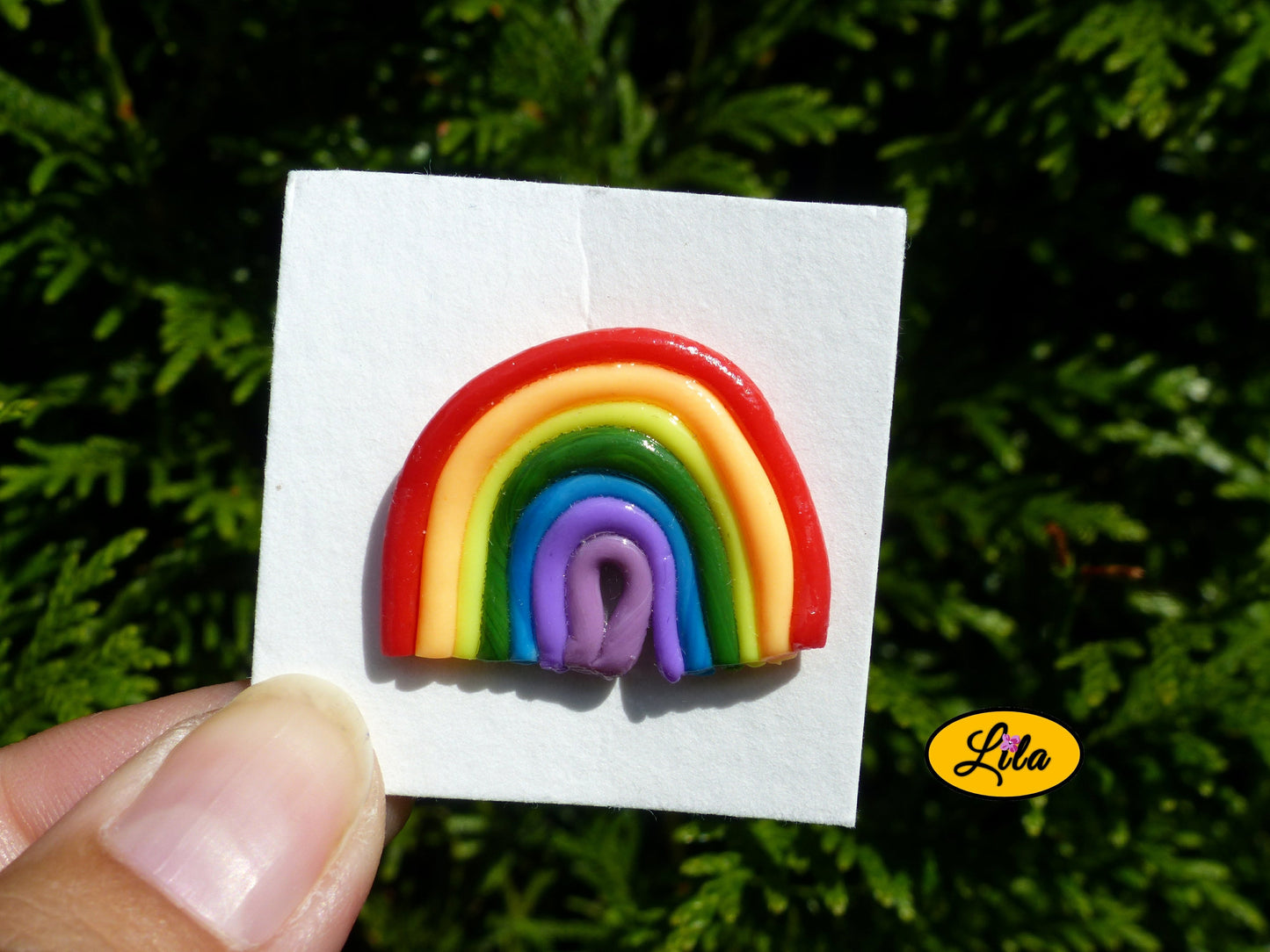 Pin’s arc-en-ciel réalisé en pâte polymère sur carton de présentation blanc photographié devant une haie style coloré et kawaii