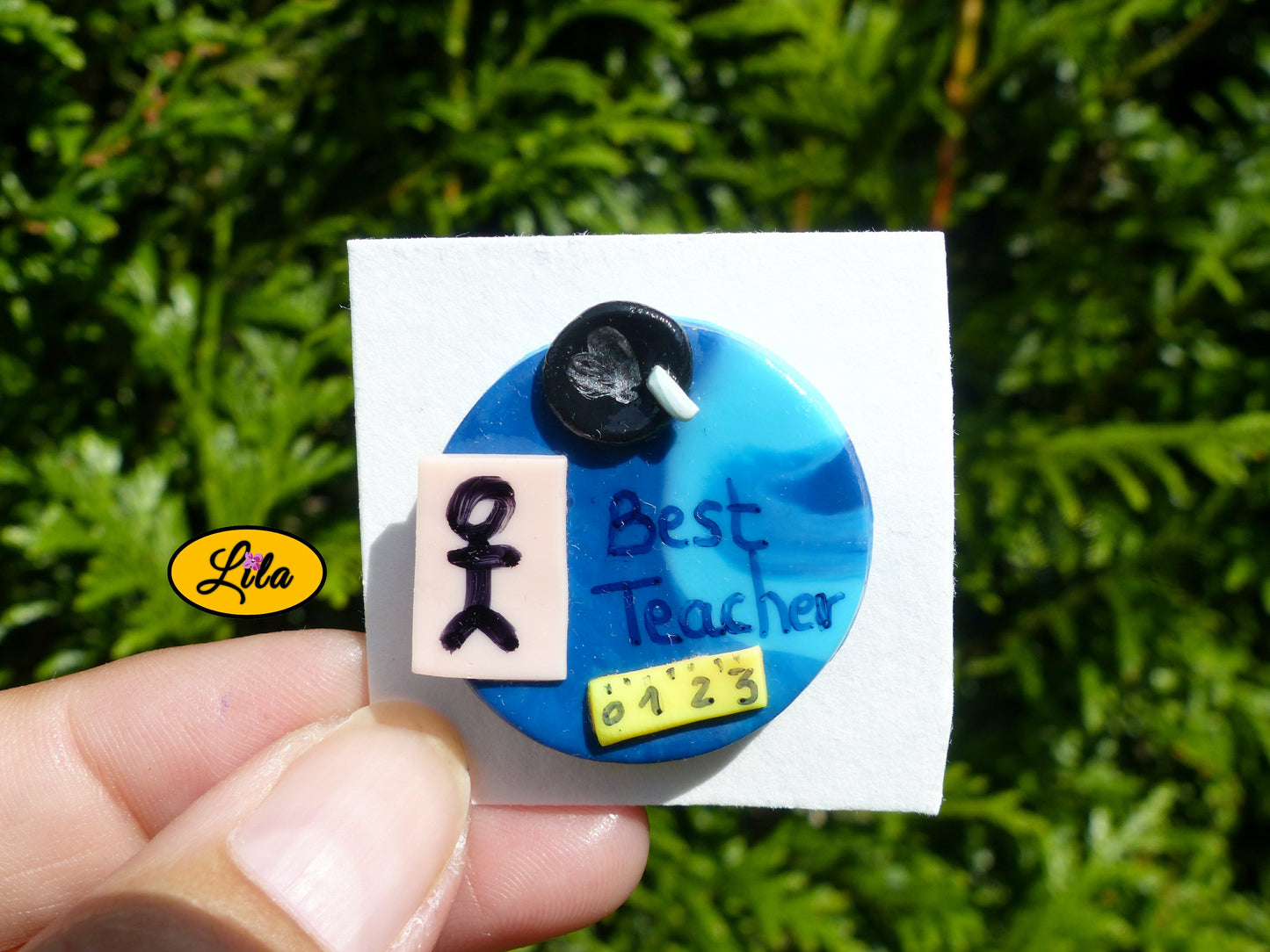 Pin’s Best Teacher bleu réalisé en pâte polymère avec effet tableau d’école, petit bonhomme décoratif, craie et inscription Best Teacher photographié devant une haie style cadeau de remerciement pour instituteur ou institutrice