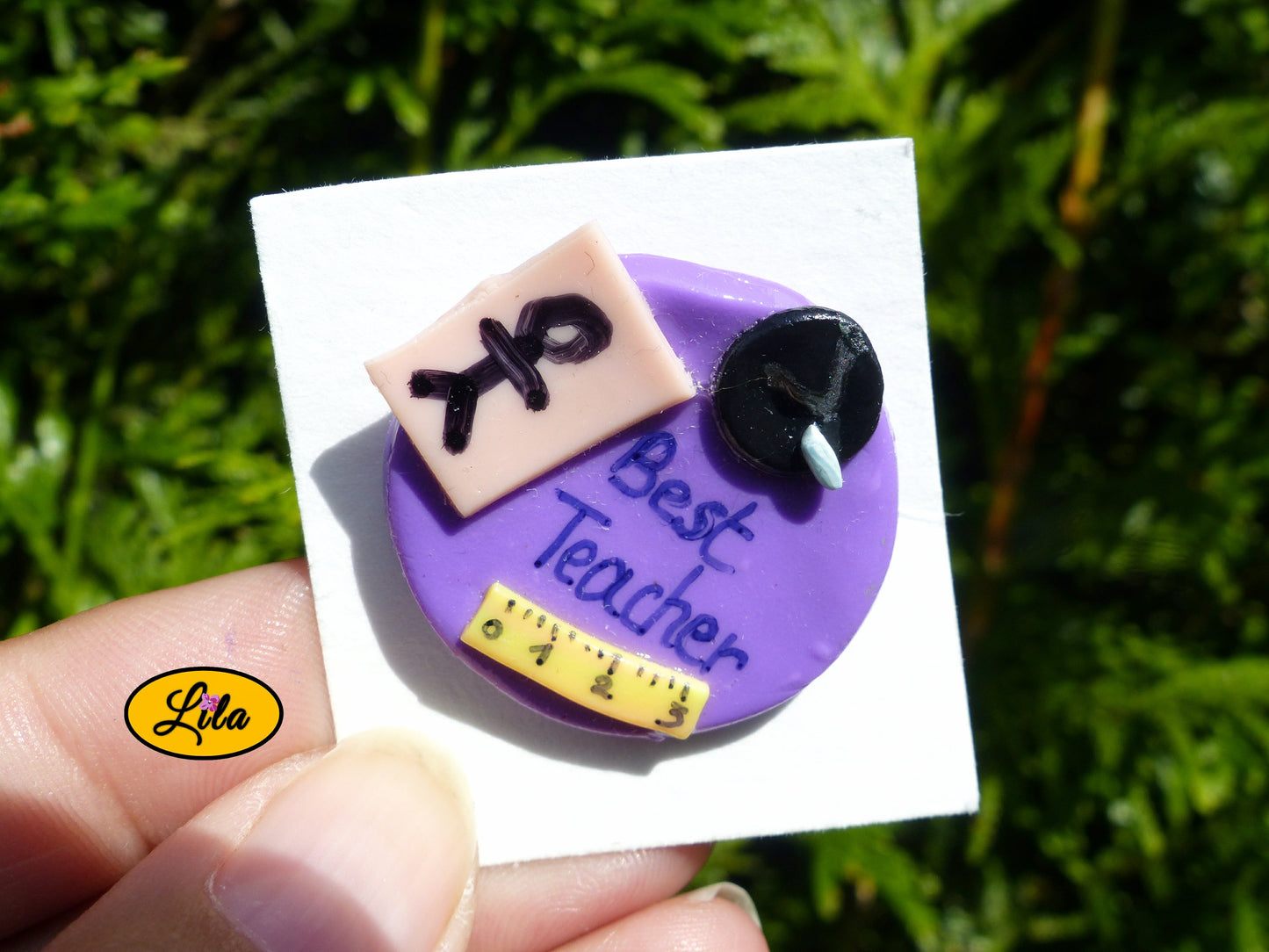 Pin’s Best Teacher mauve réalisé en pâte polymère avec effet tableau d’école, petit bonhomme décoratif, craie et inscription Best Teacher photographié devant une haie style cadeau de remerciement pour instituteur ou institutrice