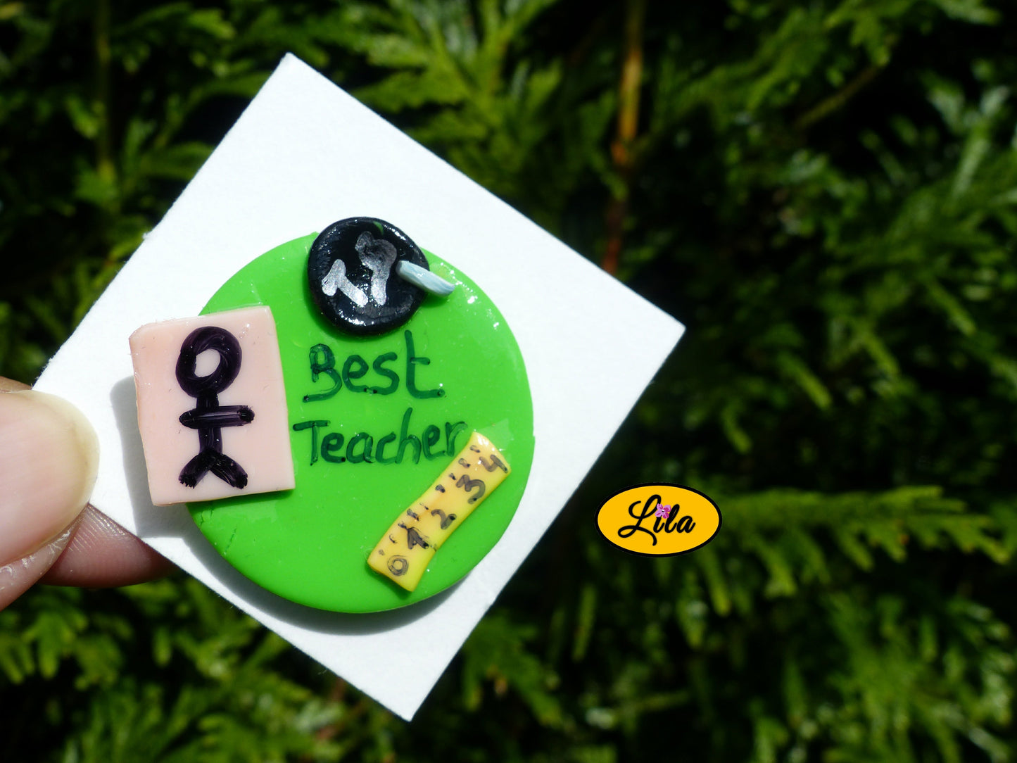 Pin’s Best Teacher réalisé en pâte polymère avec rond vert effet tableau d’école, petit bonhomme décoratif, craie et inscription Best Teacher photographié devant une haie style cadeau de remerciement pour instituteur ou institutrice