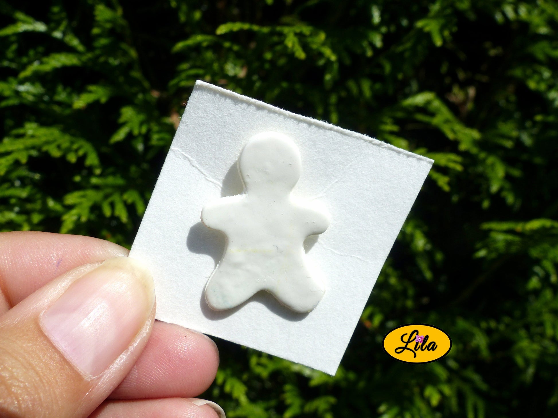 Pin’s bonhomme blanc réalisé en pâte polymère sur carton de présentation blanc photographié devant une haie style kawaii et artisanal