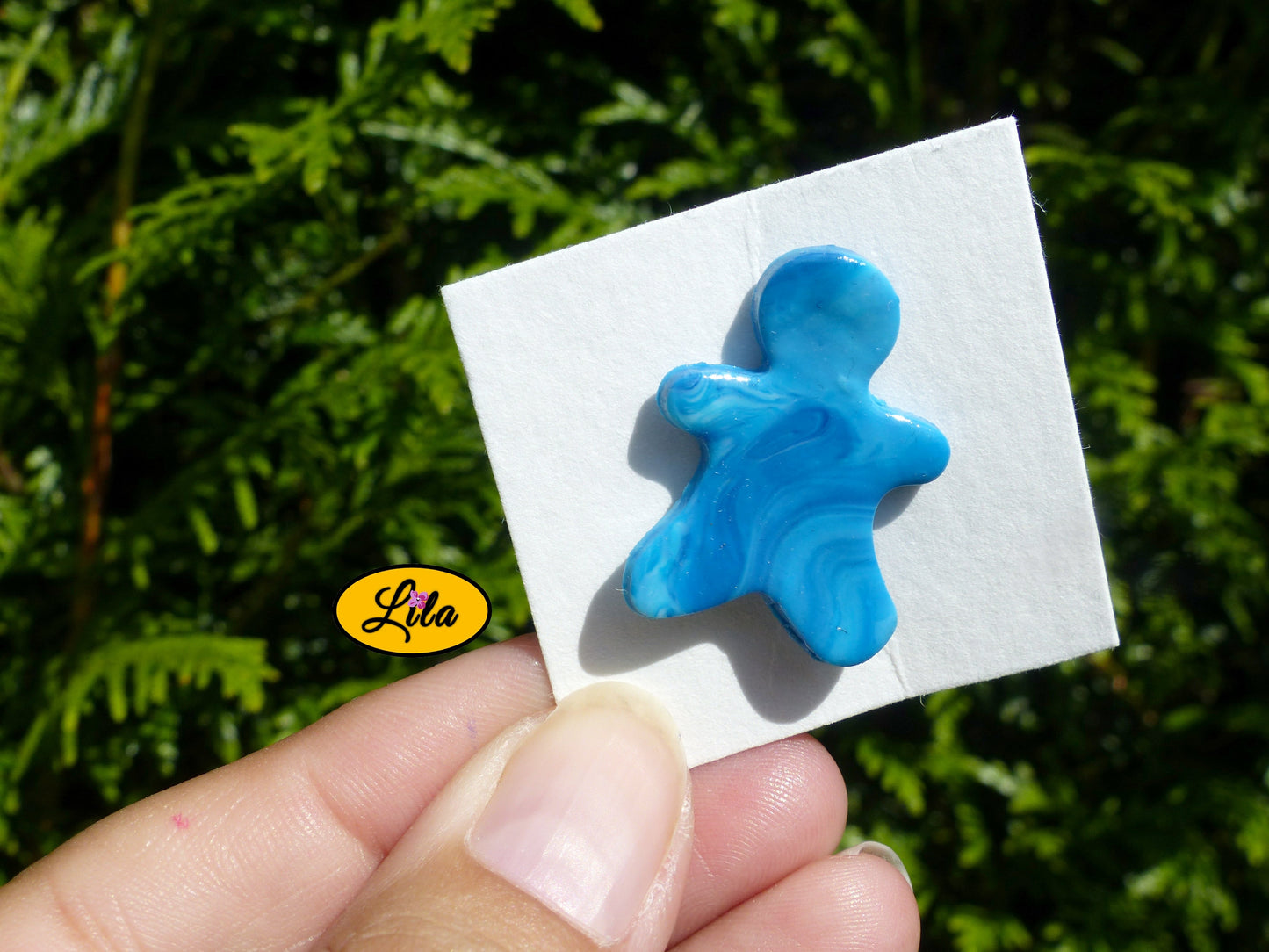 Pin’s bonhomme bleu réalisé en pâte polymère sur carton de présentation blanc photographié devant une haie style kawaii et artisanal