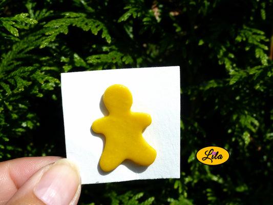 Pin’s bonhomme jaune réalisé en pâte polymère sur carton de présentation blanc photographié devant une haie style kawaii et artisanal