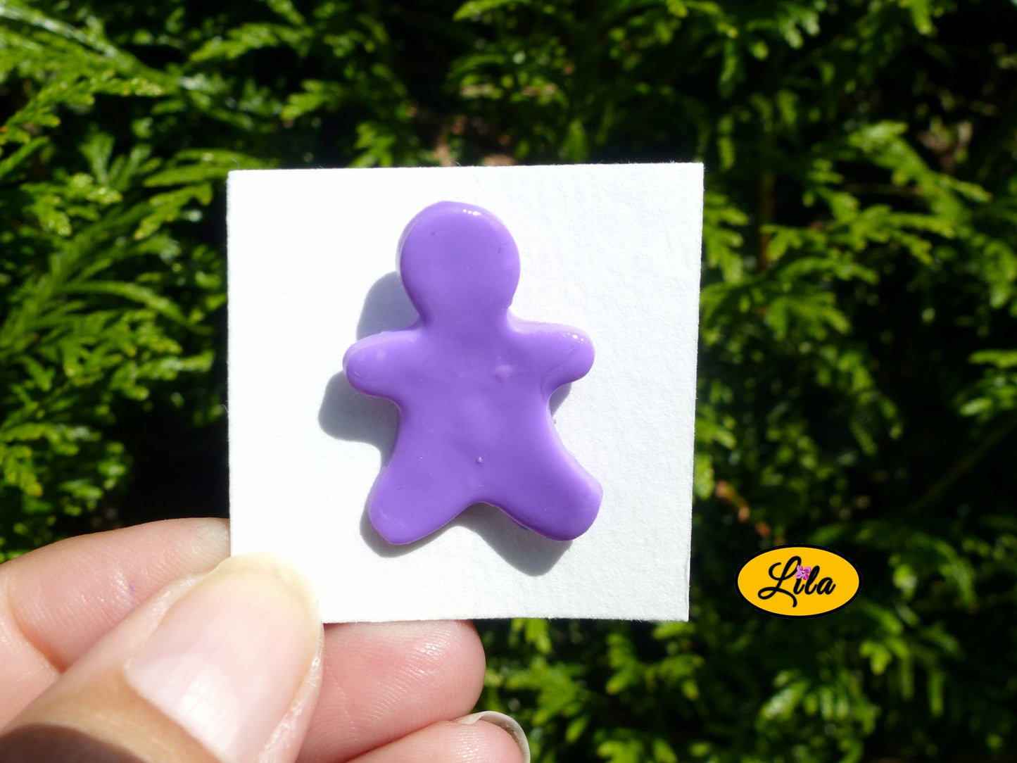 Pin’s bonhomme mauve réalisé en pâte polymère sur carton de présentation blanc photographié devant une haie style kawaii et artisanal