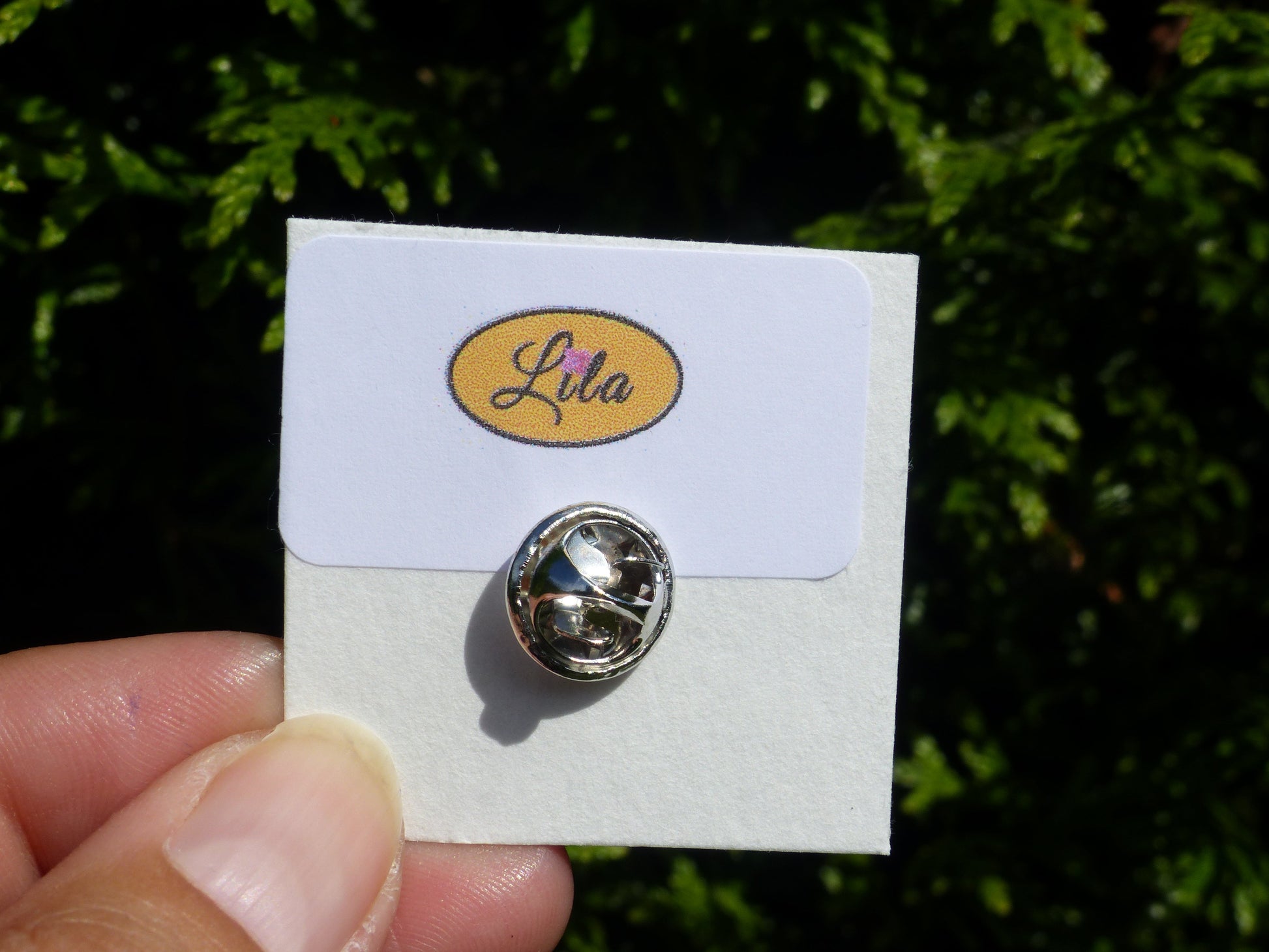 Pin’s bonhomme rose réalisé en pâte polymère avec attache argentée sur carton de présentation blanc photographié devant une haie style kawaii et artisanal