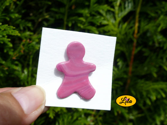 Pin’s bonhomme rose réalisé en pâte polymère sur carton de présentation blanc tenu du bout des doigts en extérieur devant une haie style kawaii et artisanal