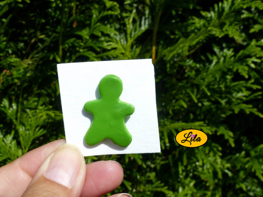 Pin’s bonhomme vert réalisé en pâte polymère sur carton de présentation blanc photographié devant une haie style kawaii et artisanal