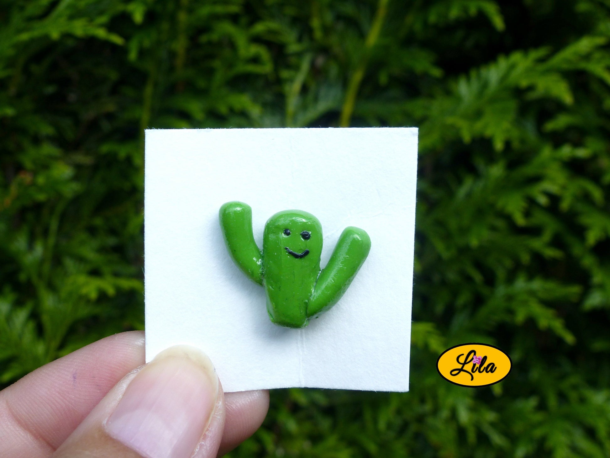 Pin’s cactus kawaii vert avec deux branches levées réalisé en pâte polymère sur carton de présentation blanc photographié devant une haie style fun et artisanal