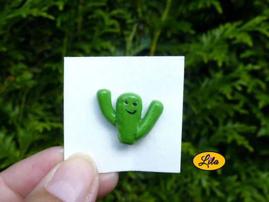 Pin’s cactus kawaii vert avec deux branches levées réalisé en pâte polymère sur carton de présentation blanc photographié devant une haie style fun et artisanal