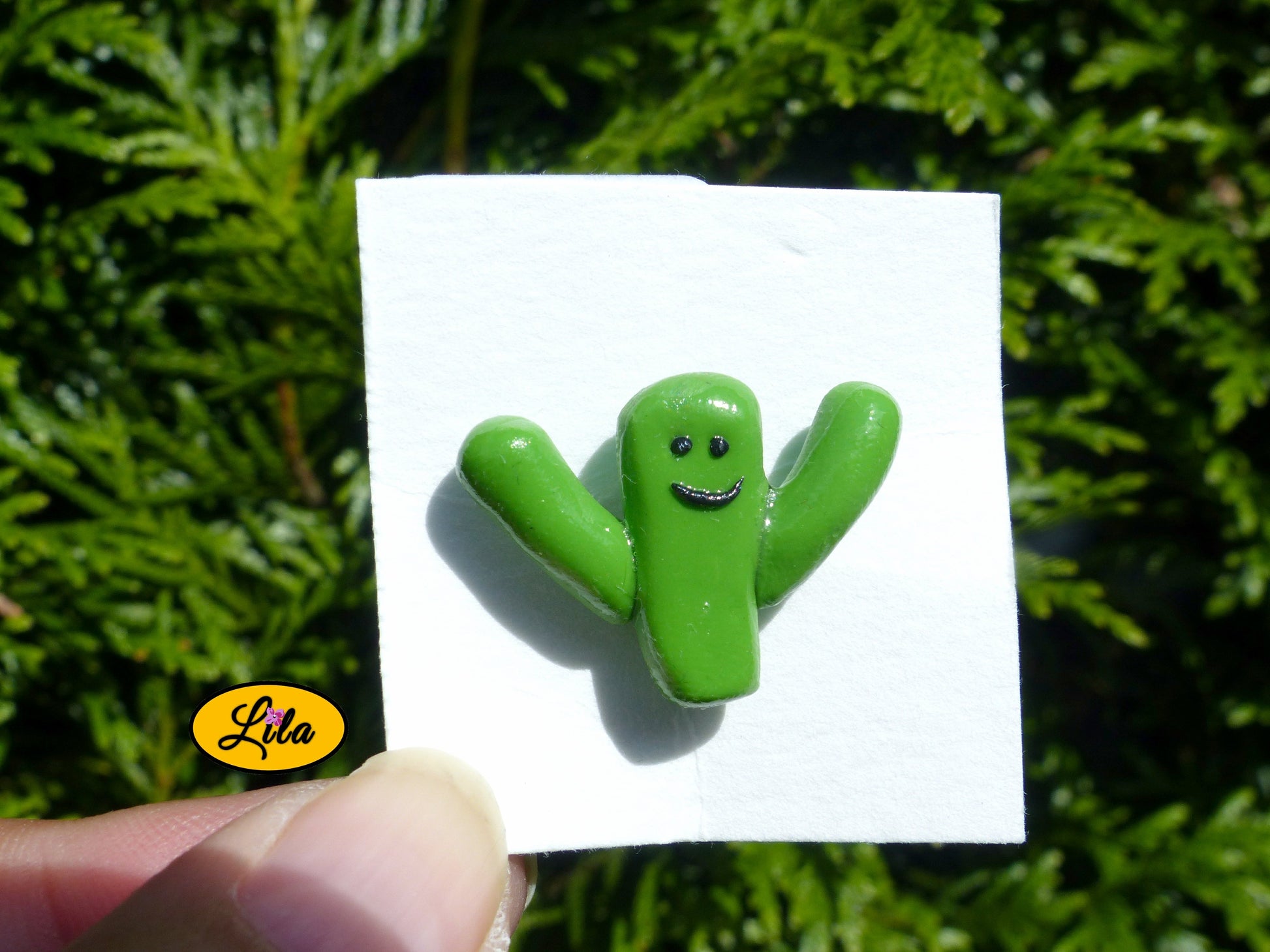 Pin’s cactus kawaii vert réalisé en pâte polymère sur carton de présentation blanc photographié devant une haie style fun et artisanal