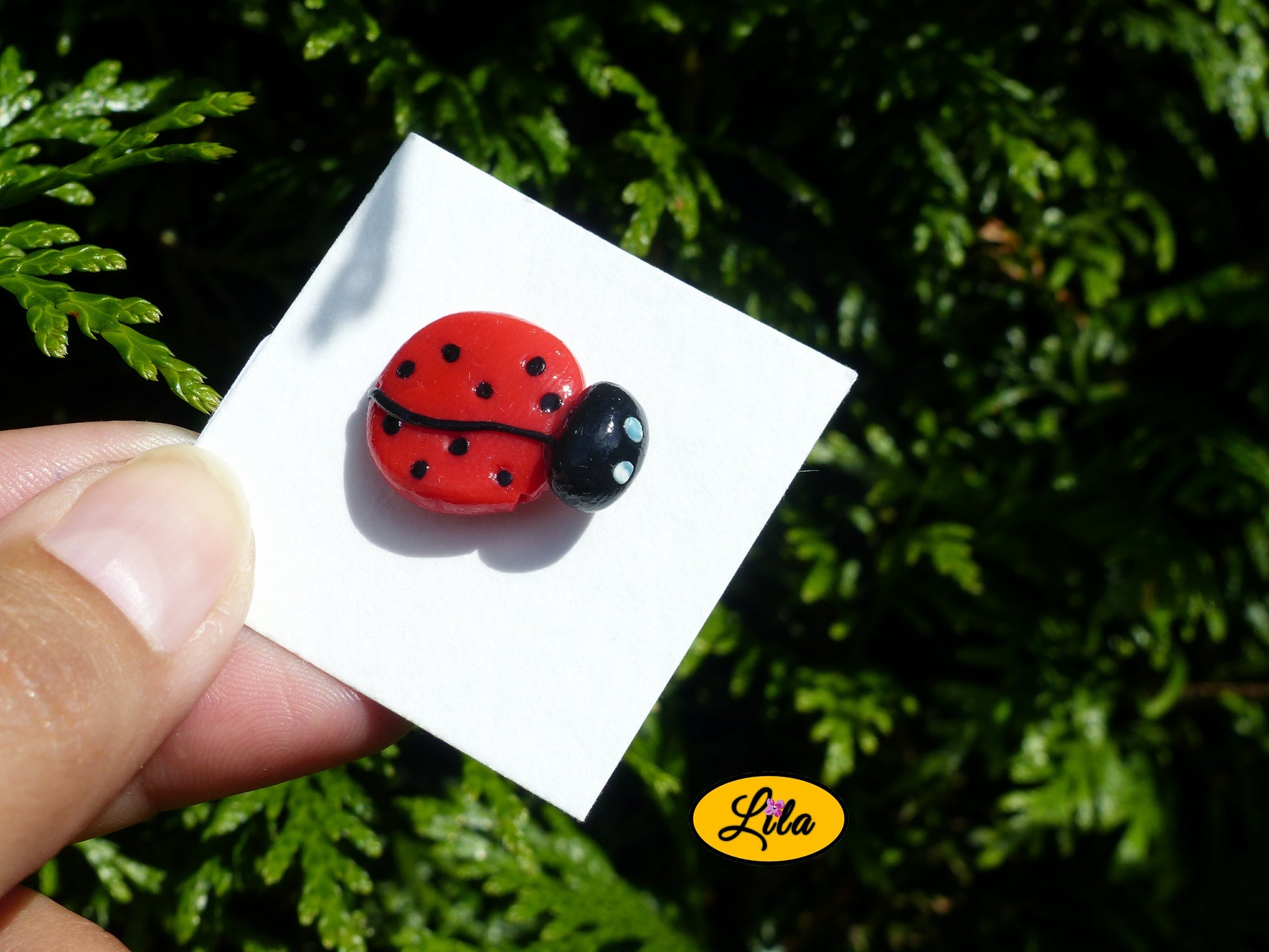 Pin’s coccinelle réalisé en pâte polymère sur carton de présentation blanc légèrement penché vers l’arrière photographié devant une haie style artisanal et kawaii