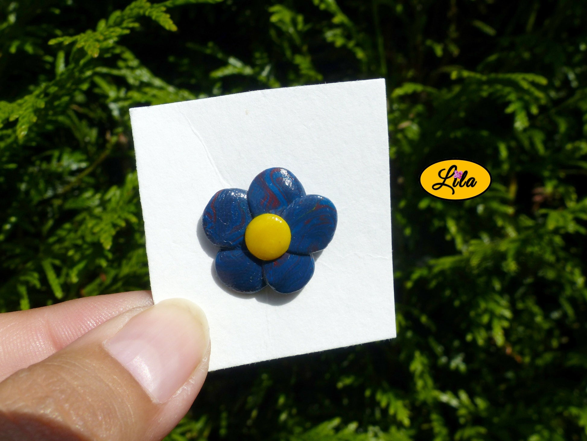 Pin’s fleur aux pétales bleu foncé avec cœur jaune réalisé en pâte polymère sur carton de présentation blanc légèrement penché vers l’arrière photographié devant une haie style floral et artisanal